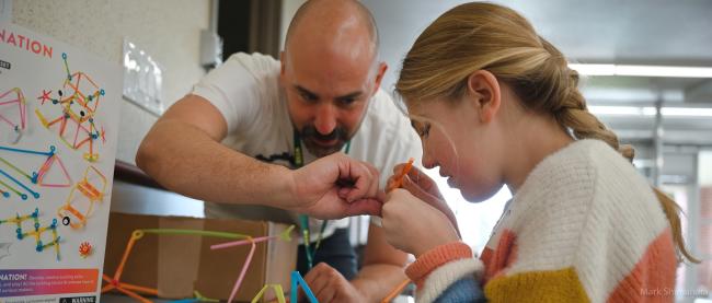 A girl with low vision closely examines elements of a 3-dimensional activity involving straws and connectors. A teacher is assisting. 