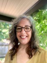 Pam Parker, an older woman with a big welcoming smile and greying hair, wearing black glasses and pale gold top.