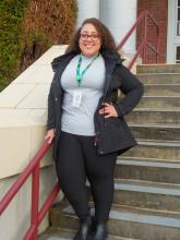 Kayla is smiling while standing posed on the stairs with her hand on her hip while holding the the stair railing.  She has really curly brown hair and wears glasses