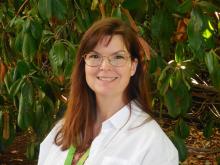 Jenny Donaldson is standing in front of a rhododendron bush while wearing a white top and green lanyard and glasses.  Jenny’s medium brown hair is highlighted by the sunshine.