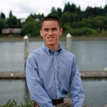 Song standing in front of the water wearing a blue button up shirt and smiling